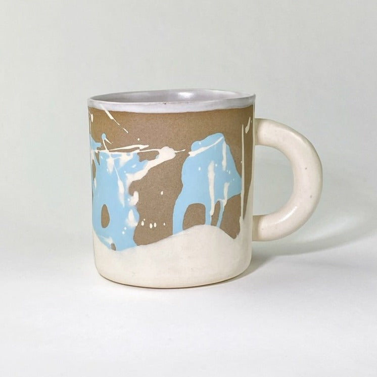 White stoneware snow like hills with desert sand skies, bright blue background with white stoneware splatters. Matte white liner on the inside and on handle